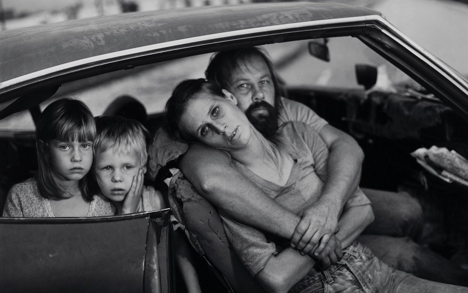 A family sat in a car with an open door. A man and woman embrace in the front seat while two children look out from the back.