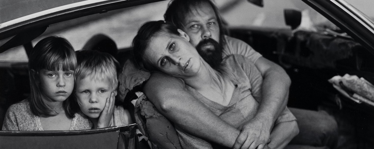 A family sat in a car with an open door. A man and woman embrace in the front seat while two children look out from the back.