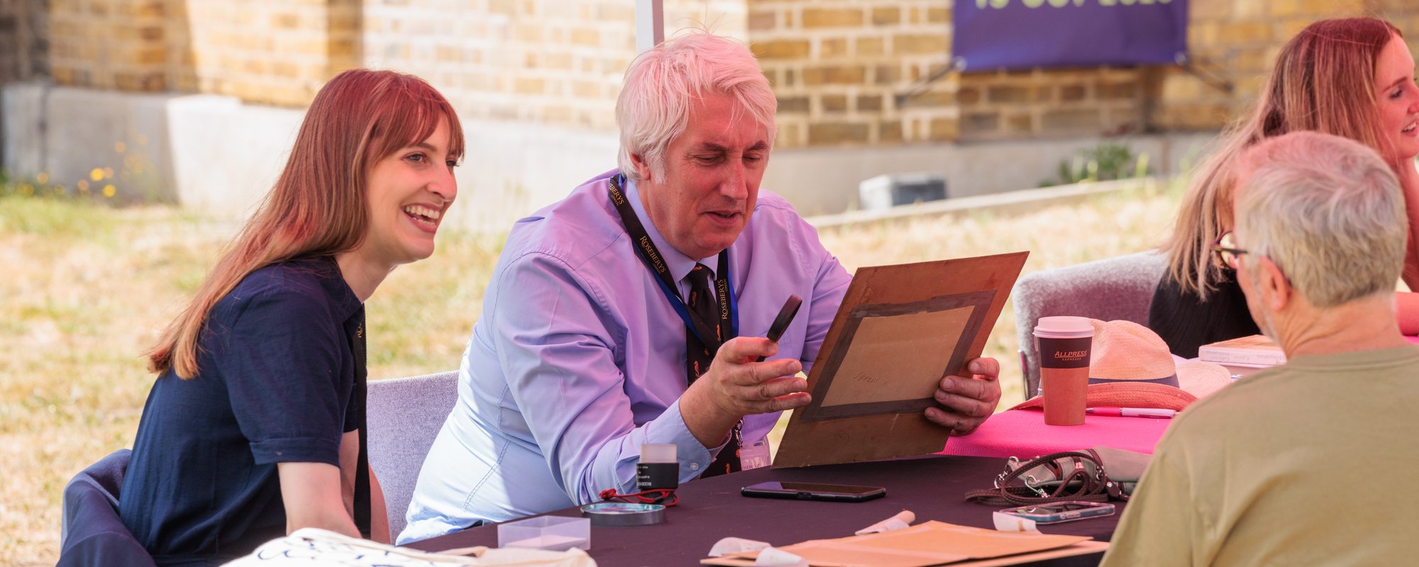 A man sat at a table outdoors holding a painting. A woman is sat next to him on the left.