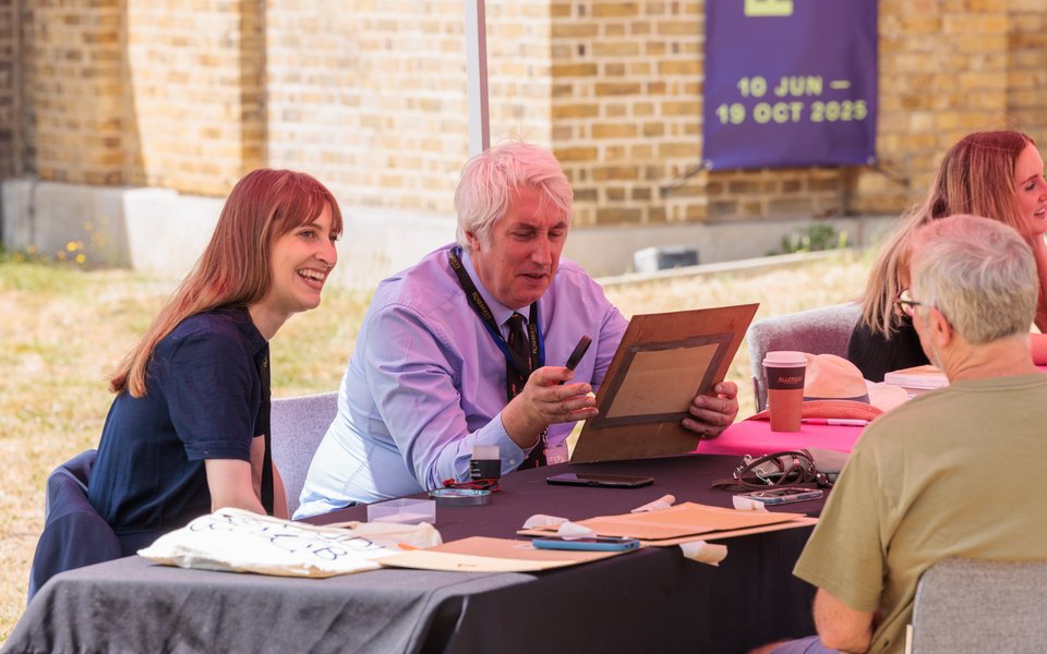 A man sat at a table outdoors holding a painting. A woman is sat next to him on the left.