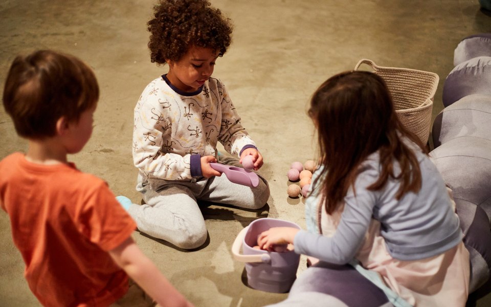 Three children sit on the floor, joyfully playing with various stuffed animals around them.
