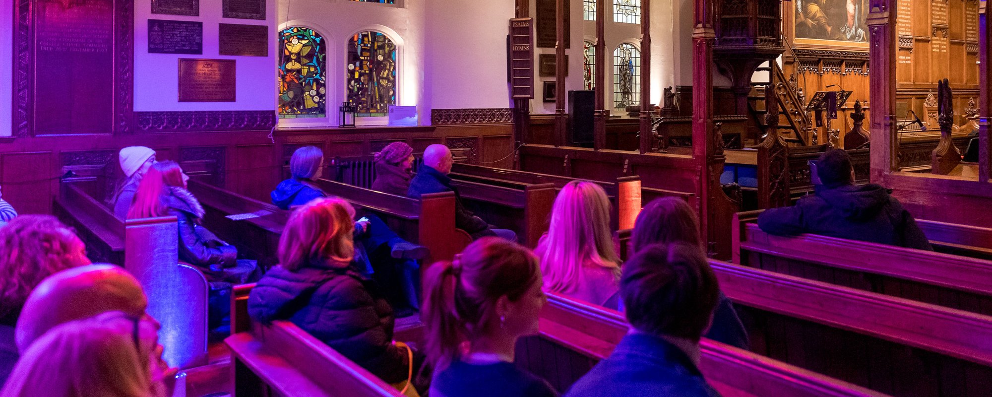 A group of people seated in a church, surrounded by colorful stained glass windows illuminating the interior.