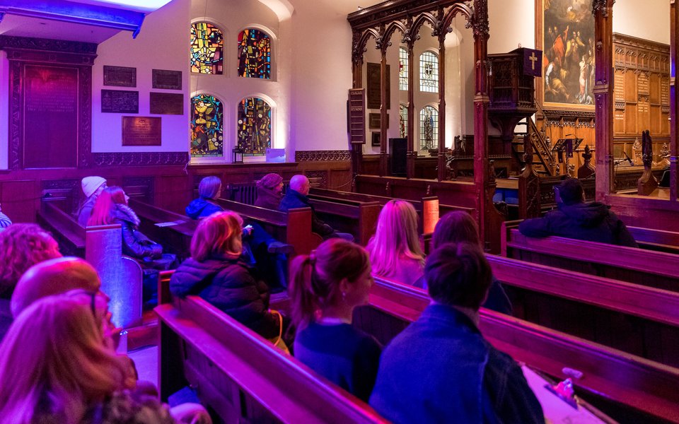 A group of people seated in a church, surrounded by colorful stained glass windows illuminating the interior.