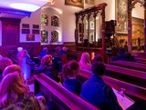A group of people seated in a church, surrounded by colorful stained glass windows illuminating the interior.