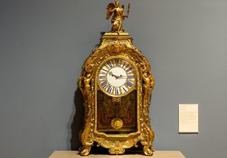 An elaborate gold clock with a glass door showing a swinging sun pendulum with gold statuettes on top.
