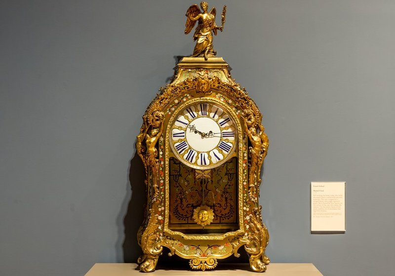 An elaborate gold clock with a glass door showing a swinging sun pendulum with gold statuettes on top.