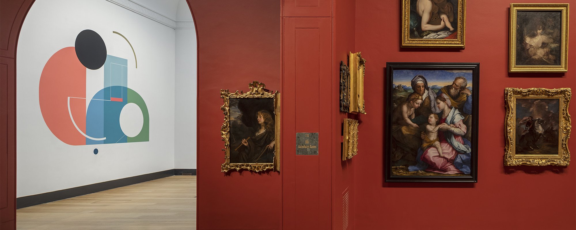 A view of the interior of Dulwich Picture Gallery. Through an arch you can see the white walls and an abstract mural of the entrance hall and the main gallery space next to it with red walls and gold framed historic paintings.