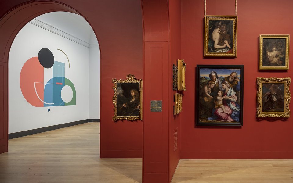 A view of the interior of Dulwich Picture Gallery. Through an arch you can see the white walls and an abstract mural of the entrance hall and the main gallery space next to it with red walls and gold framed historic paintings.