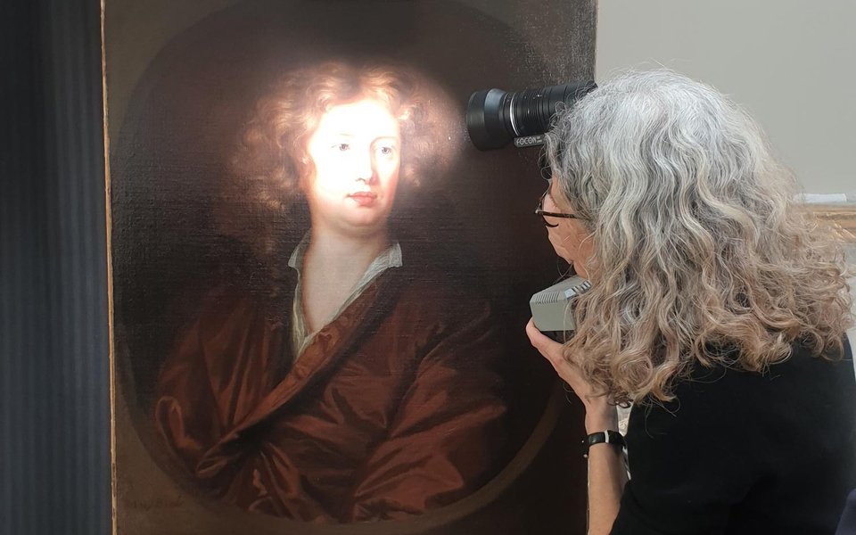 A woman looks closely at a portrait with a torch. The painting sits on an easel.