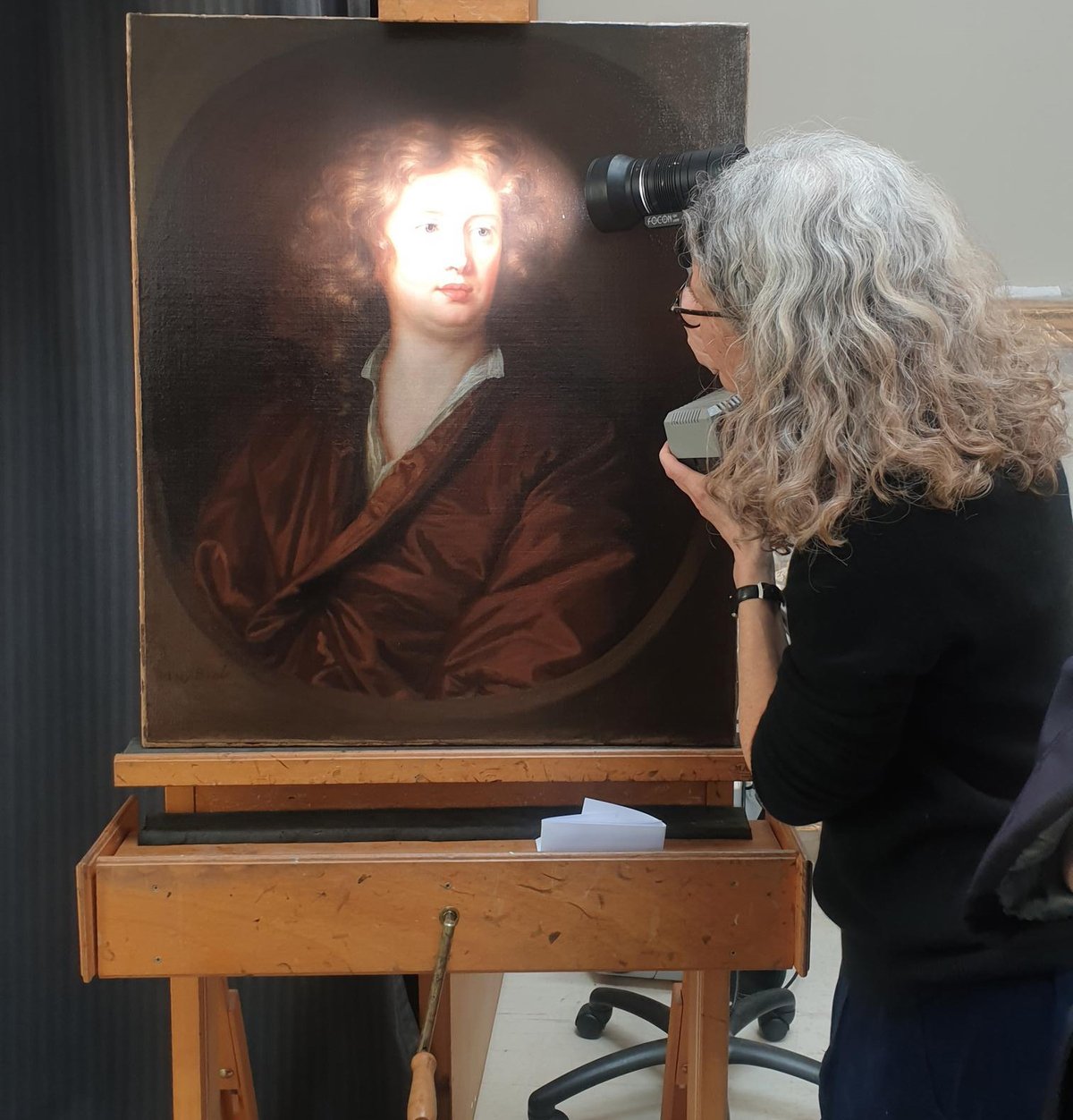 A woman looks closely at a portrait with a torch. The painting sits on an easel.