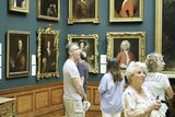 People observe framed portraits in an art gallery with blue walls and wooden paneling. Some individuals stand closely examining the artworks.