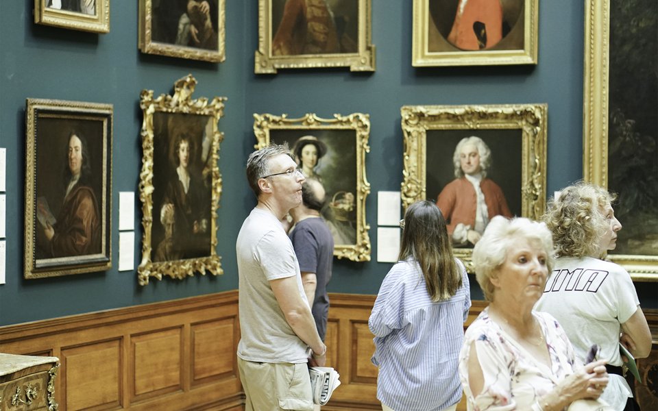 People observe framed portraits in an art gallery with blue walls and wooden paneling. Some individuals stand closely examining the artworks.