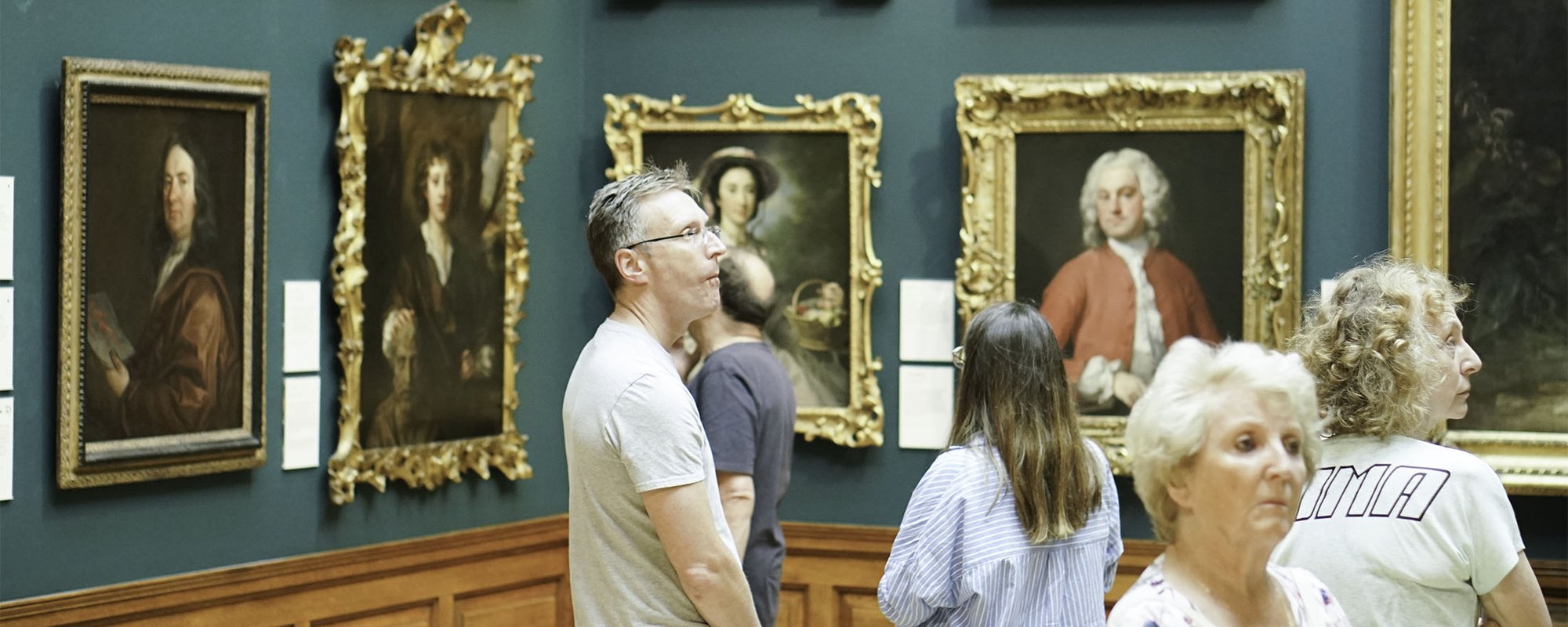 People observe framed portraits in an art gallery with blue walls and wooden paneling. Some individuals stand closely examining the artworks.