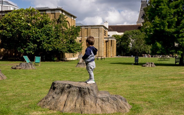 Child standing on a bronze stump in a field next to a brick building