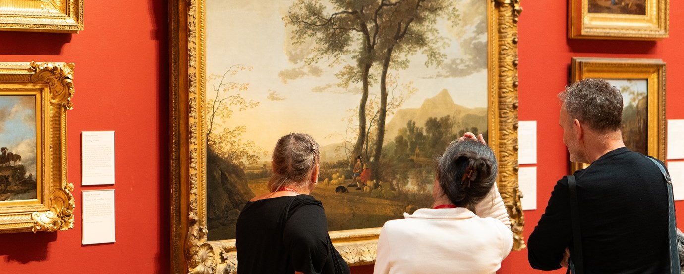 A group of three people stand looking at a large landscape painting in a gold frame hung on a red wall.