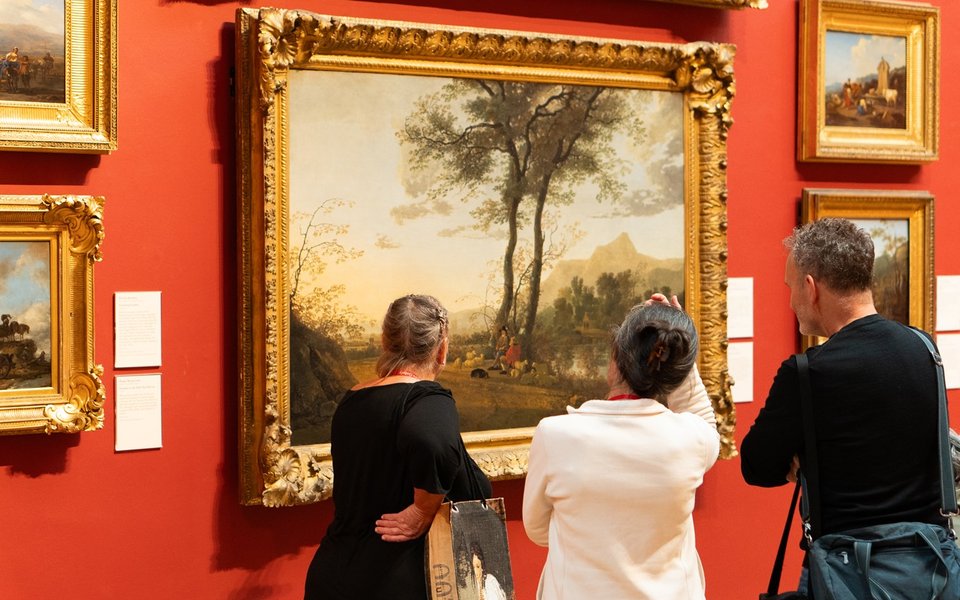 A group of three people stand looking at a large landscape painting in a gold frame hung on a red wall.