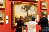 A group of three people stand looking at a large landscape painting in a gold frame hung on a red wall.
