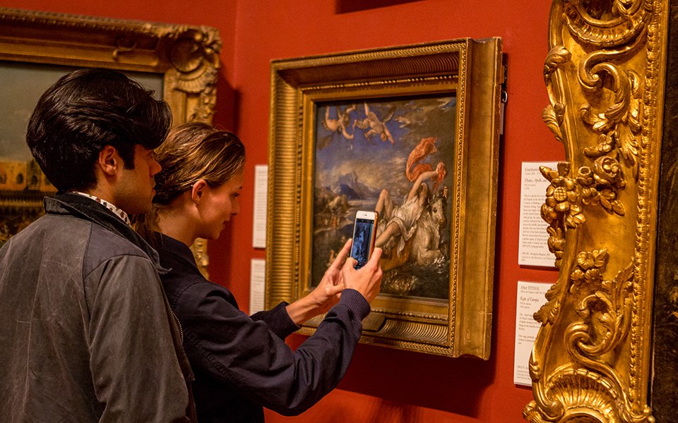 man and a woman stand in front of a gold framed painting. The woman holds her phone up to the artwork.