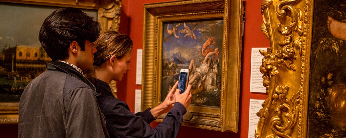 man and a woman stand in front of a gold framed painting. The woman holds her phone up to the artwork.