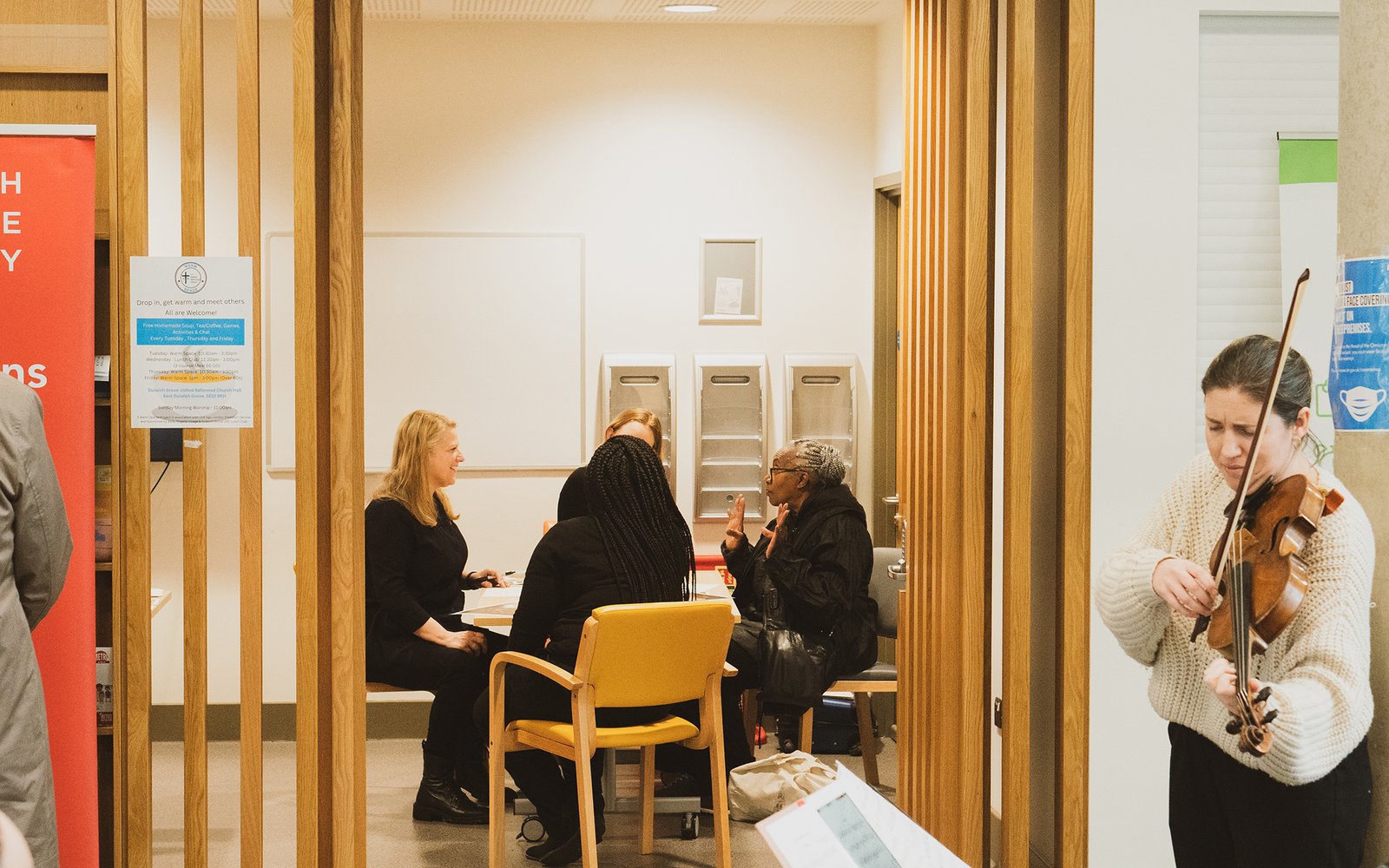 A woman stands playing a violin outside a doorway through which you can see a small group of people sat at a table chatting.