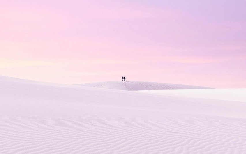 A couple walking on a large dune with sunset sky behind.